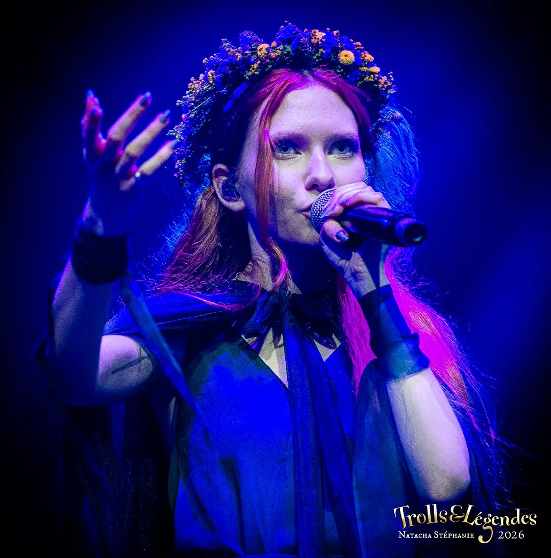 LILITH MAX chante sur scène lors du Festival 2026 (Hub: Concerts). under une lumière bleue électrique, elle porte une couronne de fleurs, des cheveux roses, une cape sombre et des gants, micro en main, énergie envoûtante.