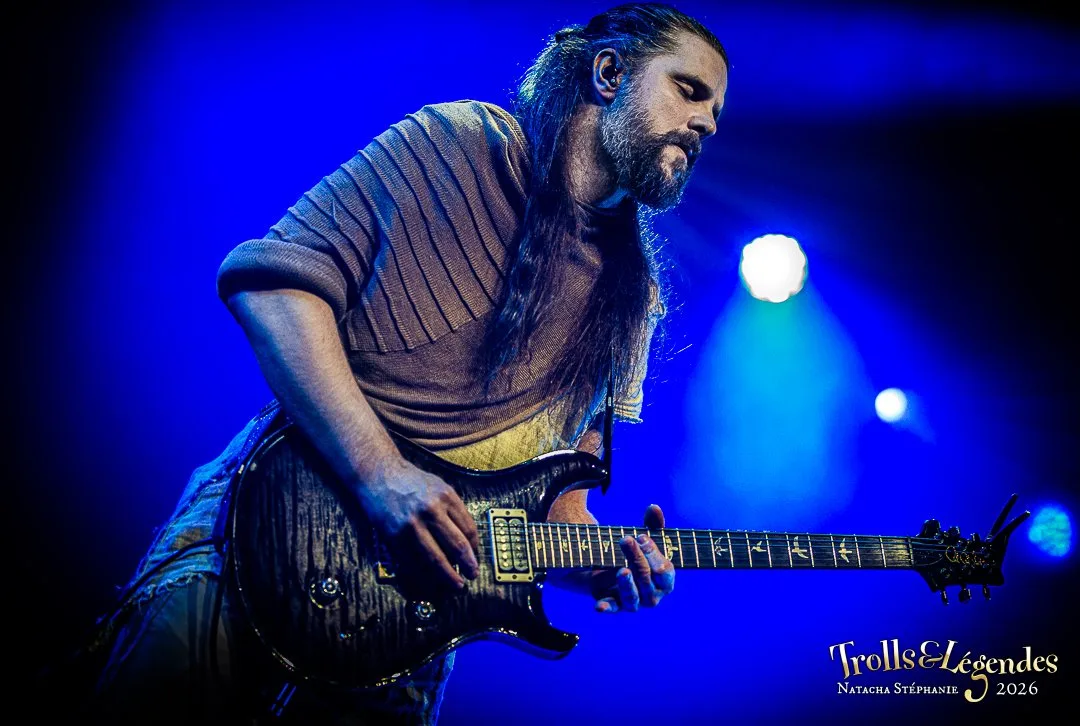 Kalandra sur scène au hub Concerts, Festival 2026. Guitare électrique sombre, jeu précis, long cheveux noués et barbe. Lumières bleues enveloppantes, ambiance intense et mystique, riffs captivants et énergie rock.