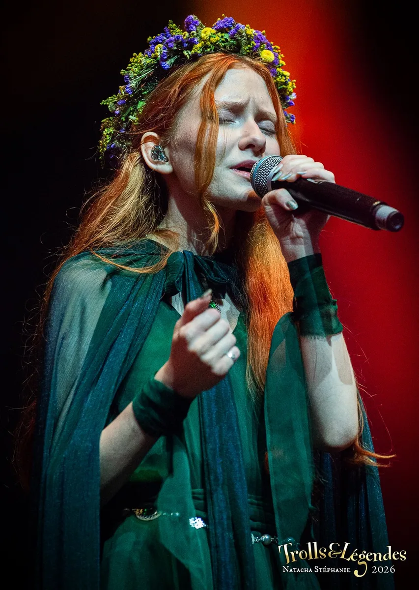 LILITH MAX chante avec intensité sur scène, couronne de fleurs, longes cheveux rouges et cape vert émeraude. Micro en main, yeux fermés, énergie féerique dans une lumière rouge. Hub: Concerts, Événement: Festival 2026.