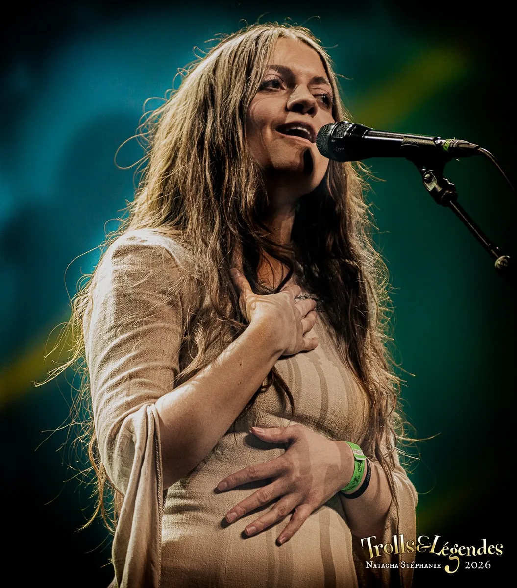 Kalandra chante sur scène au festival 2026, hub Concerts. Elle pose une main sur la poitrine, l’autre près du micro, expression émue, lumière chaude et fond coloré qui accentue l’intimité du moment.