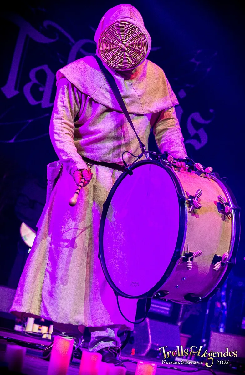 TABERNIS sur scène au Festival 2026 (Hub: Concerts). Musicien enveloppé d'une robe beige, masque rond tissé couvrant le visage, jouant d’un grand tambour circulaire accroché devant lui. Lumières violettes et ambiance immersive.