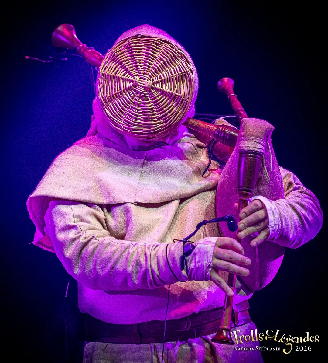 TABERNIS sur scène au Festival 2026 (Hub: Concerts). Un musicien enveloppé d’une tunique beige porte un masque tressé en osier qui cache son visage et joue de la cornemuse sous un éclairage violet intense.