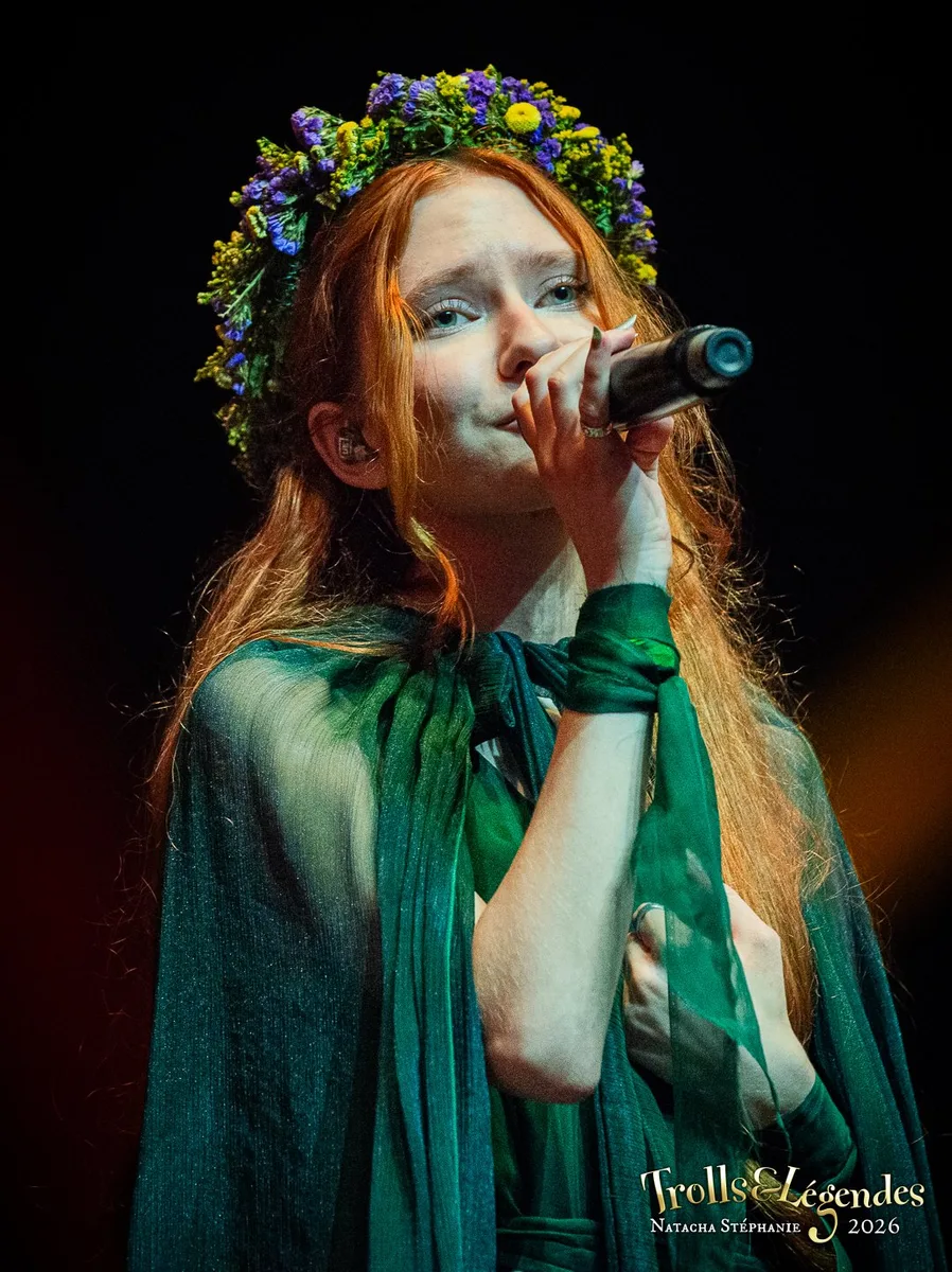 Lilith Max, chanteuse au look féerique: couronne florale violette et jaune, robe vert émeraude fluide et voile sombre. Elle chante dans un micro sous un éclairage chaleureux, sur scène lors du Festival 2026 dans le hub Concerts.