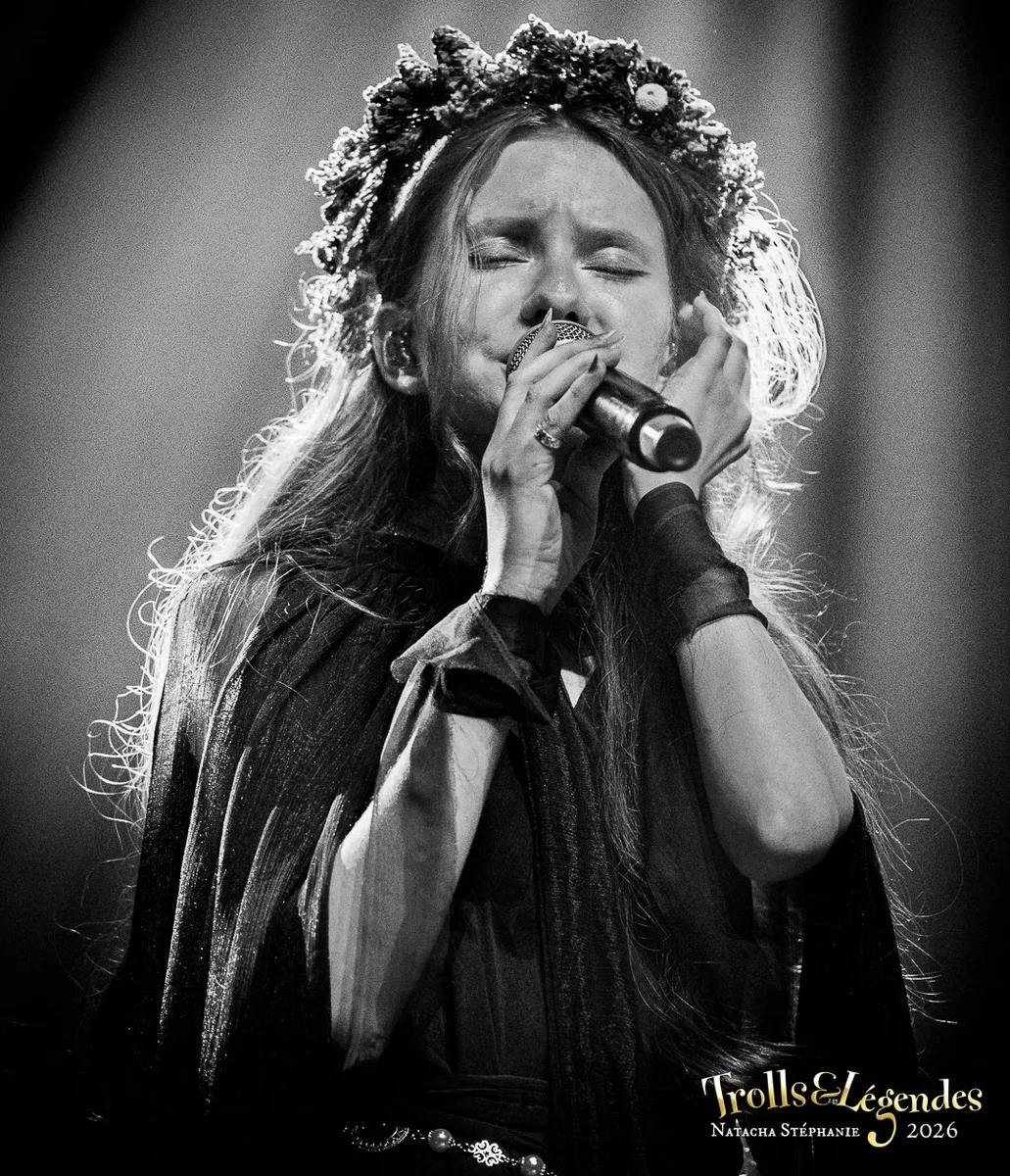 LILITH MAX chante avec intensité, couronne de fleurs et cape sombre. Yeux fermés, micro serré, énergie dramatique sur scène. Hub: Concerts; Événement: Festival 2026.