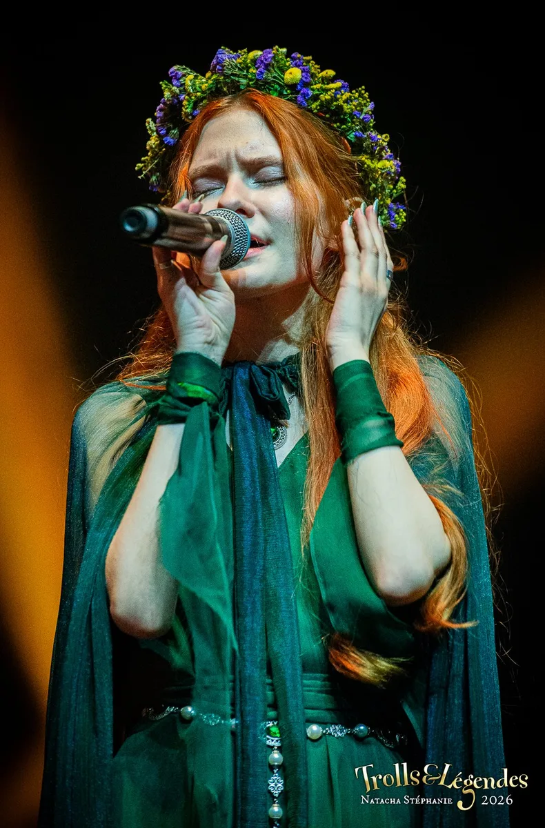 LILITH MAX en concert: chanteuse au regard intensément expressif, coiffure rouge et couronne de fleurs, vêtue d’une robe vert émeraude et d’un voile sombre. Micro à la main, elle rend la scène émouvante, dans l’ambiance du Festival 2026.