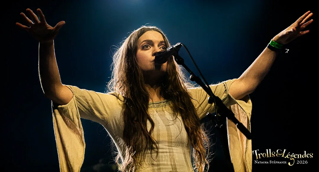 KALANDRA, chanteuse aux longs cheveux, ouvre les bras devant le micro sur scène. Robe légère, lumière dramatique et énergie vive. Présence magnétique dans le hub Concerts, during le Festival 2026.
