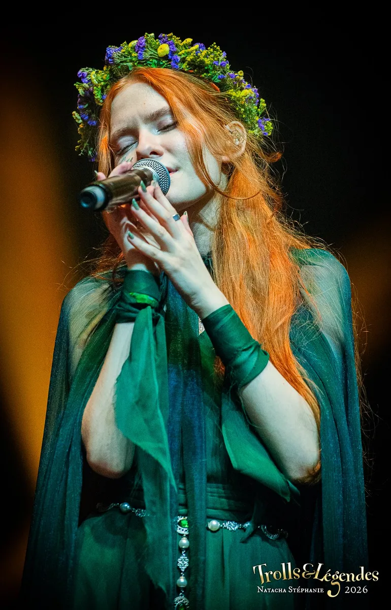 LILITH MAX sur scène, robe verte et cape translucide, couronne de fleurs violettes et jaune pâle. Elle chante les yeux clos dans le microphone, ambiance féérique et intime. Hub: Concerts. Événement: Festival 2026.