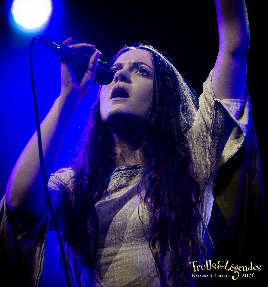 Kalandra, jeune chanteuse, prend le micro sous une lumière bleue intense, bras levés, visage exprimant passion et énergie. Scène de Concerts pour le Festival 2026, ambiance envoûtante et live.