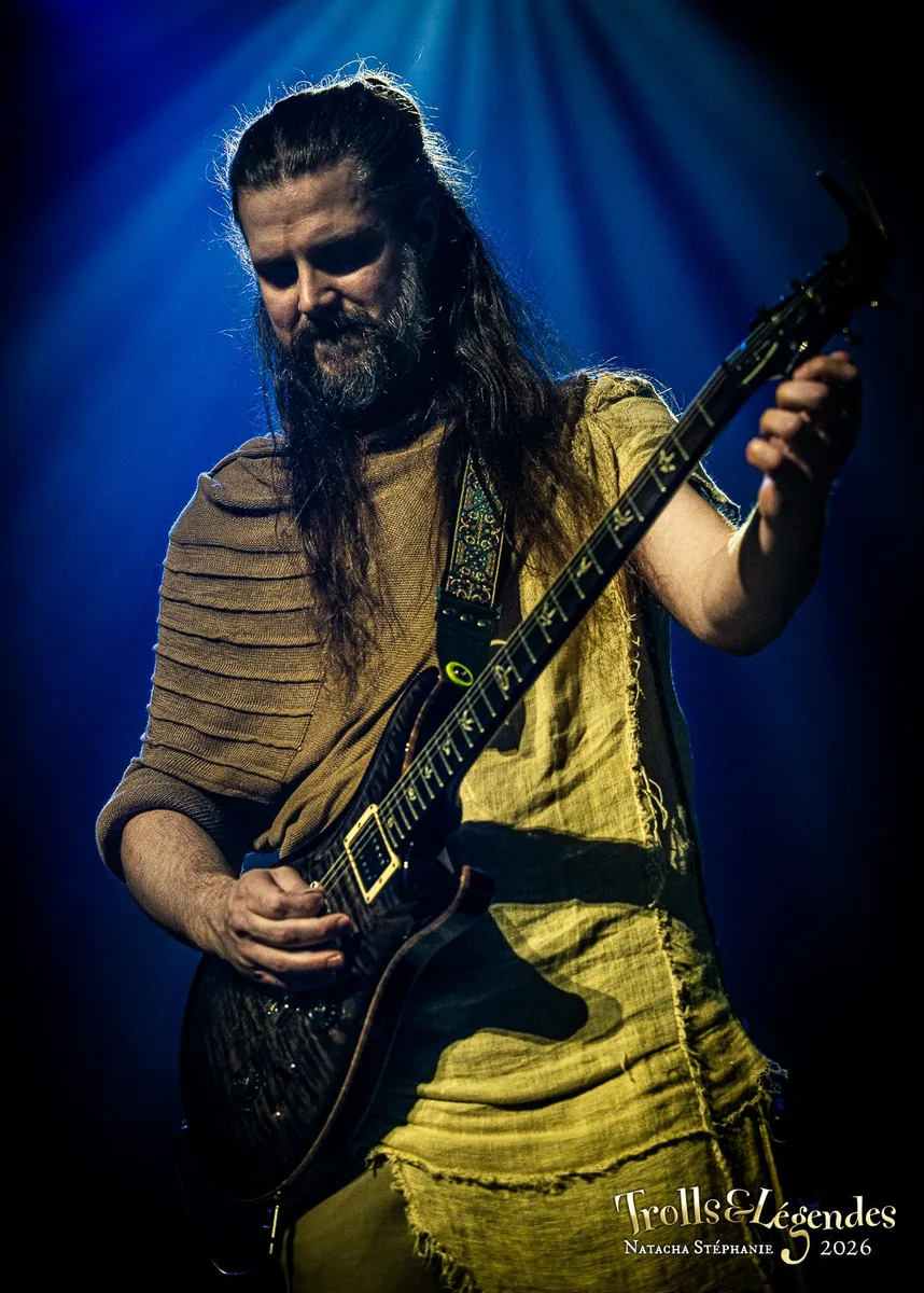Kalandra, guitariste passionné, sous lumière bleue intense. Longs cheveux et barbe, tunique beige froissée, joue une guitare électrique percussive. Concentation visible, scène sombre du Hub: Concerts, lors du Festival 2026.
